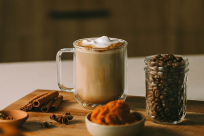 A pumpkin spice latte in a glass mug next to loose coffee beans and pumpkin puree on a countertop