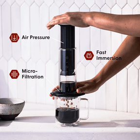 Person using an AeroPress Premium coffee maker with text labels for air pressure, fast immersion, and micro-filtration. #color_black