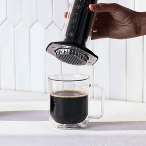 Hand using a black AeroPress Premium coffee maker to brew coffee into a glass mug against a white tiled wall. #color_black