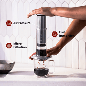 Person using a  AeroPress Premium coffee maker with text labels on air pressure, fast immersion, and micro-filtration. #color_silver