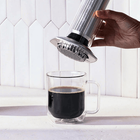 Hand using an AeroPress Premium coffee maker to pour coffee into a clear glass mug on a white background. #color_silver