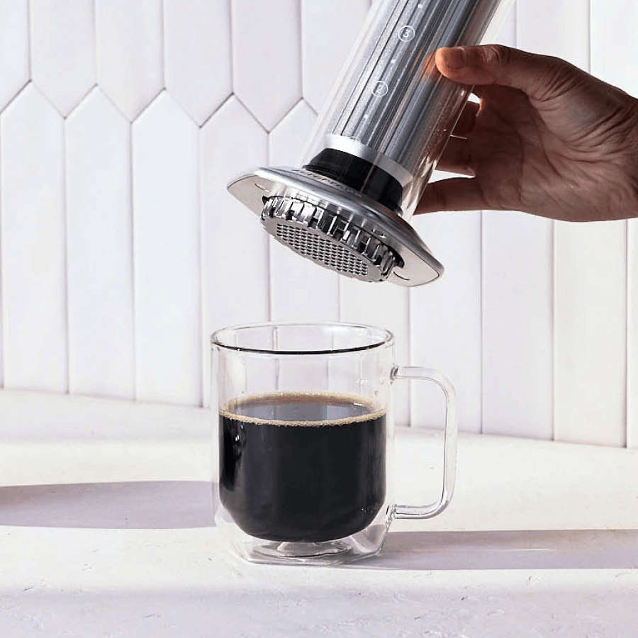 Hand using an AeroPress Premium coffee maker to pour coffee into a clear glass mug on a white background. #color_silver