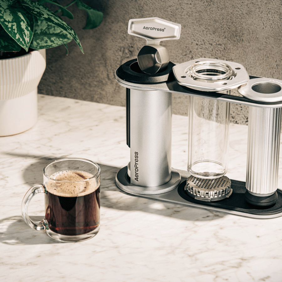 AeroPress Premium coffee maker on a marble surface with a glass of coffee next to it. #color_silver