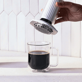 Hand using a white AeroPress Premium coffee maker to pour coffee into a clear glass mug on a white countertop. #color_white