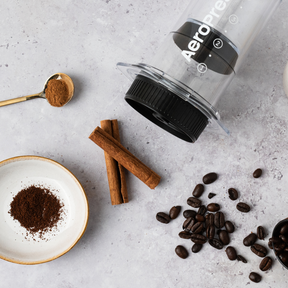 AeroPress coffee maker with coffee beans, ground coffee, and cinnamon sticks on a light gray surface. #color_black