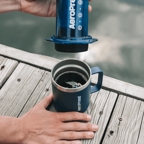 Person using an AeroPress to make coffee on a wooden deck by a body of water. #color_blue