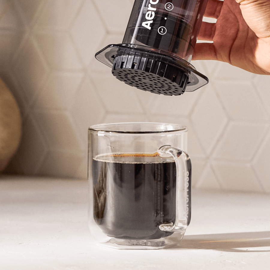 A person using an AeroPress to make coffee in a clear glass mug. #color_black