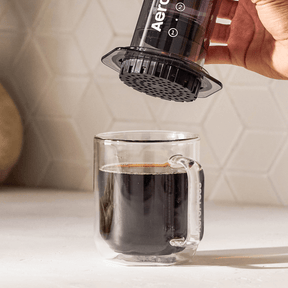 A person using an AeroPress to make coffee in a clear glass mug. #color_black
