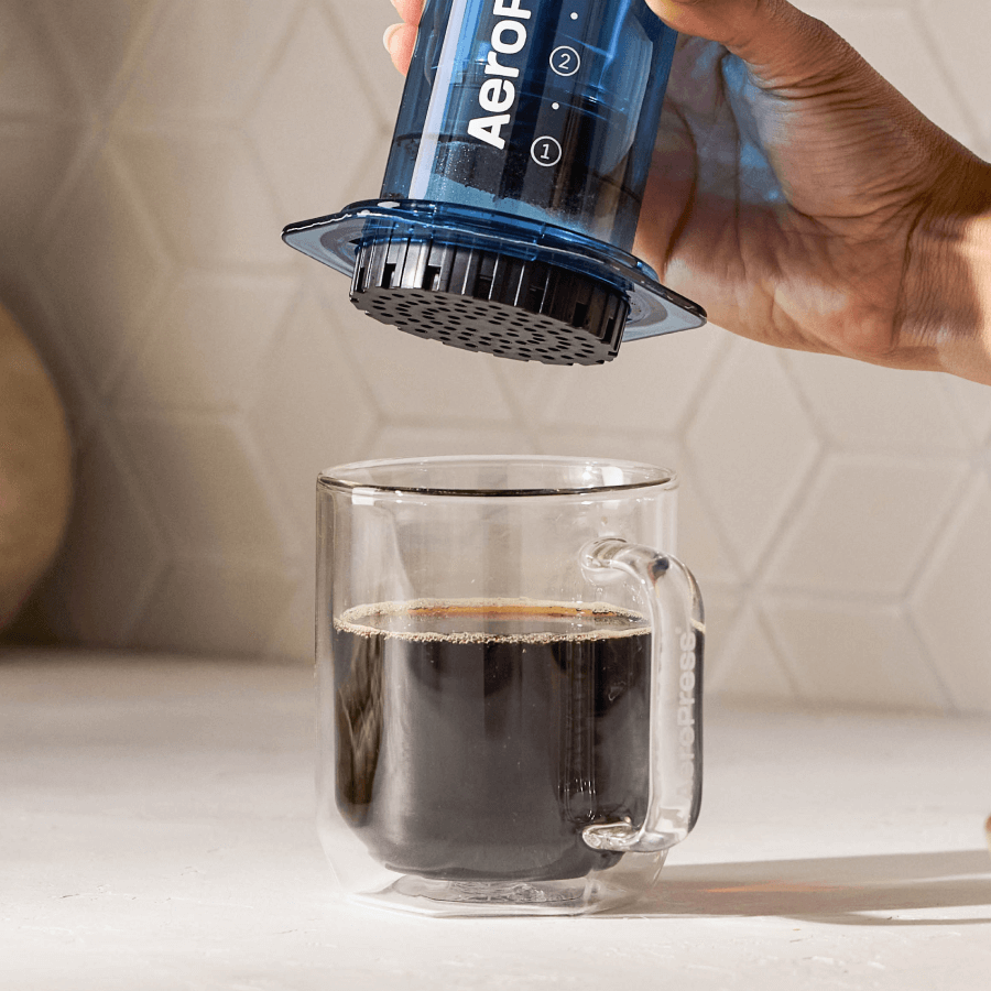 A person using an Aeropress coffee maker to pour coffee into a clear glass mug.  #color_blue