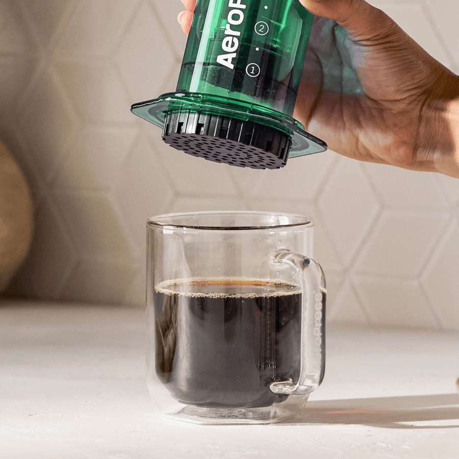 Hand holding a green AeroPress coffee maker above a glass of coffee. #color_green