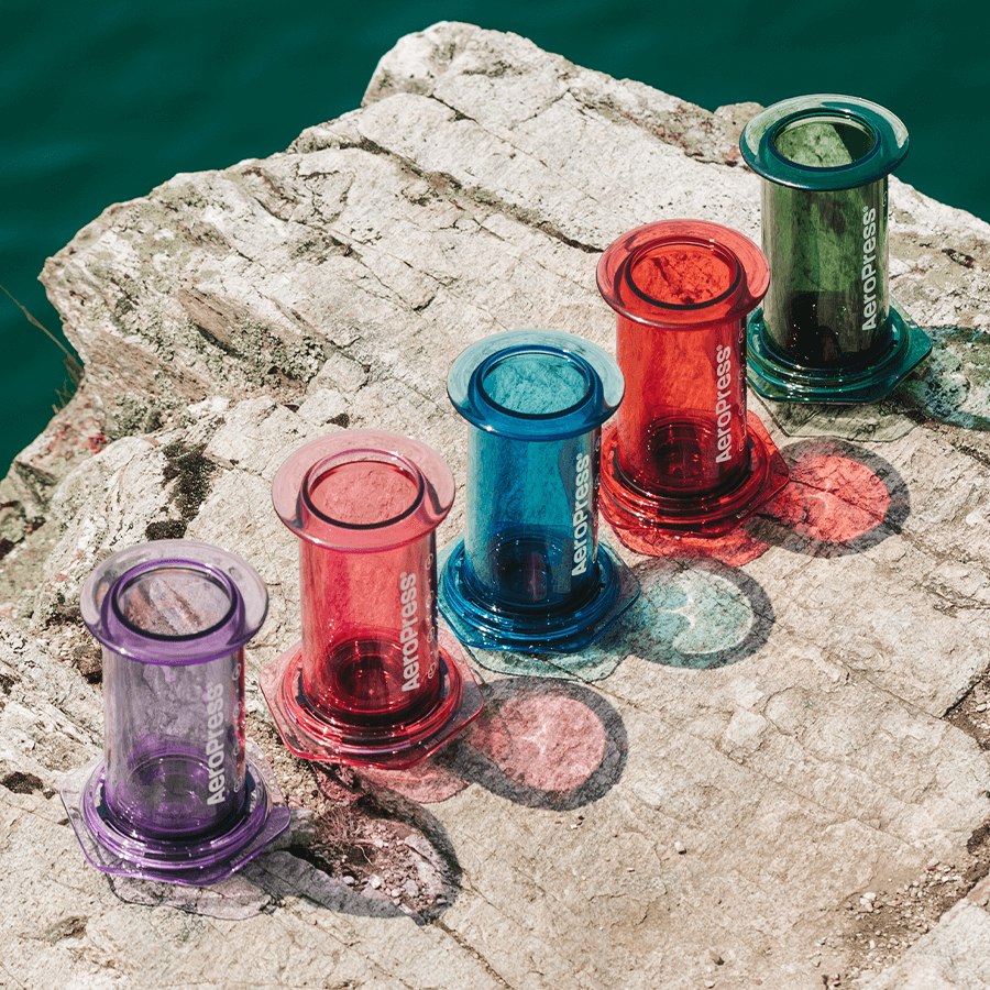 Colorful AeroPress coffee brewing devices on a rock by a body of water.