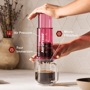 Person using a red Aeropress coffee maker with labeled features on a kitchen counter. #color_pink
