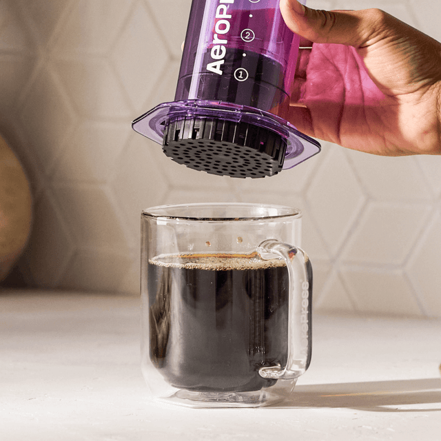 Hand using an Aeropress to make coffee in a clear glass mug. #color_purple