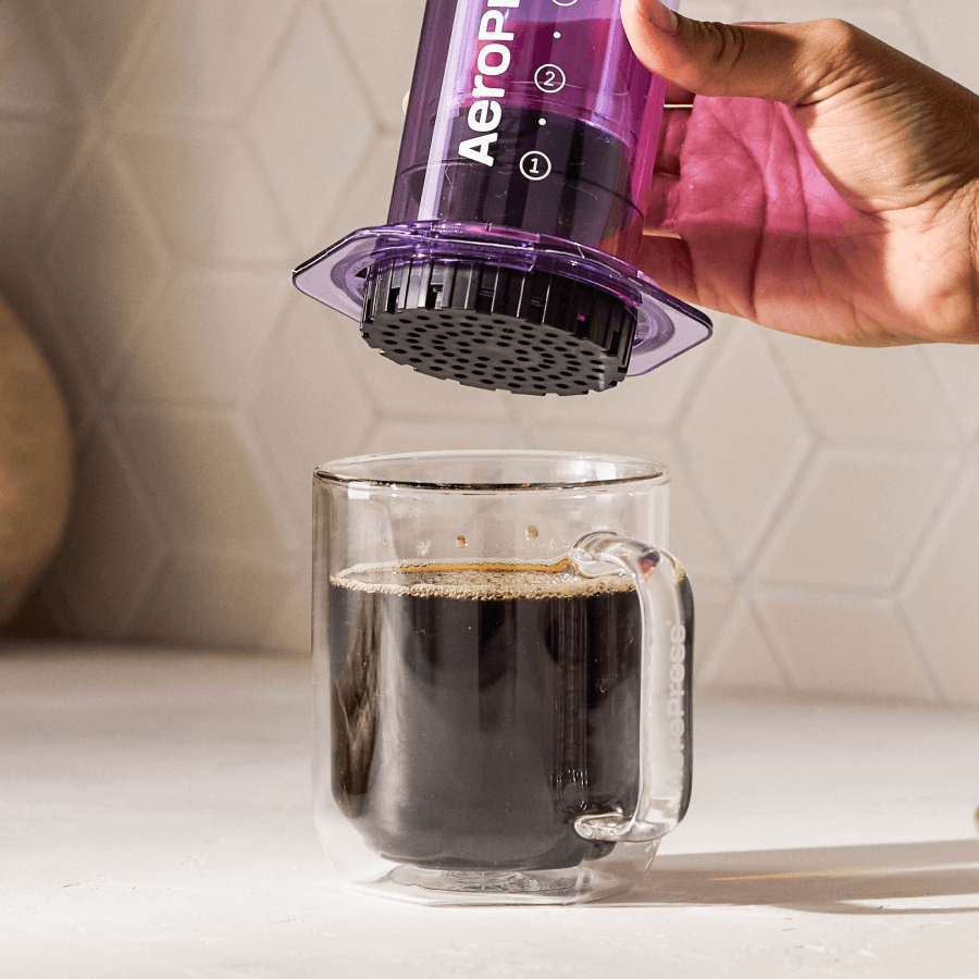 Hand using an Aeropress to make coffee in a clear glass mug. #color_purple