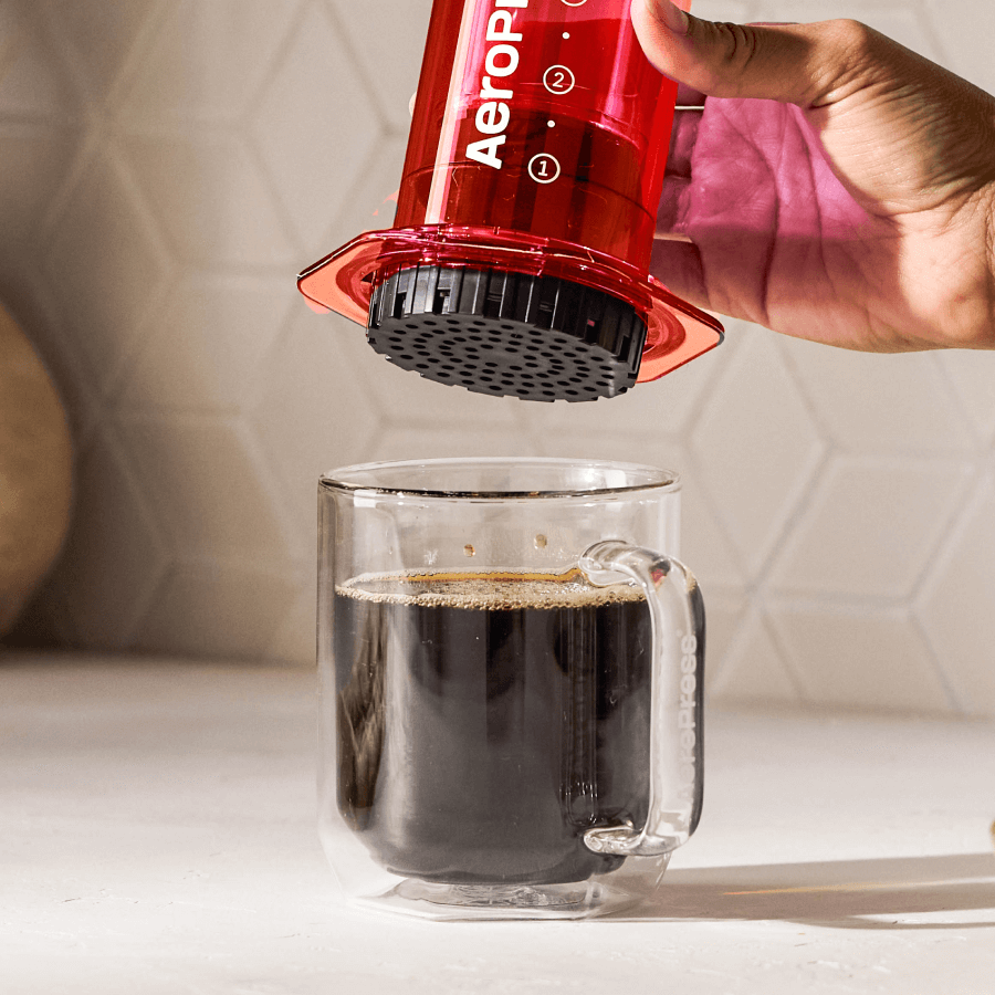 Red AeroPress device being used to make coffee in a clear glass mug. #color_red