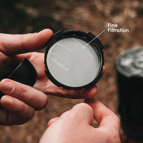 Person holding an AeroPress coffee filter with 'Fine Filtration' text, blurred natural background #color_pink