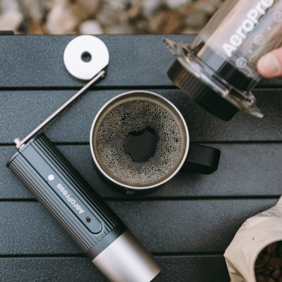 A portable AeroPress Clear coffee maker and a cup of coffee on a wooden surface.