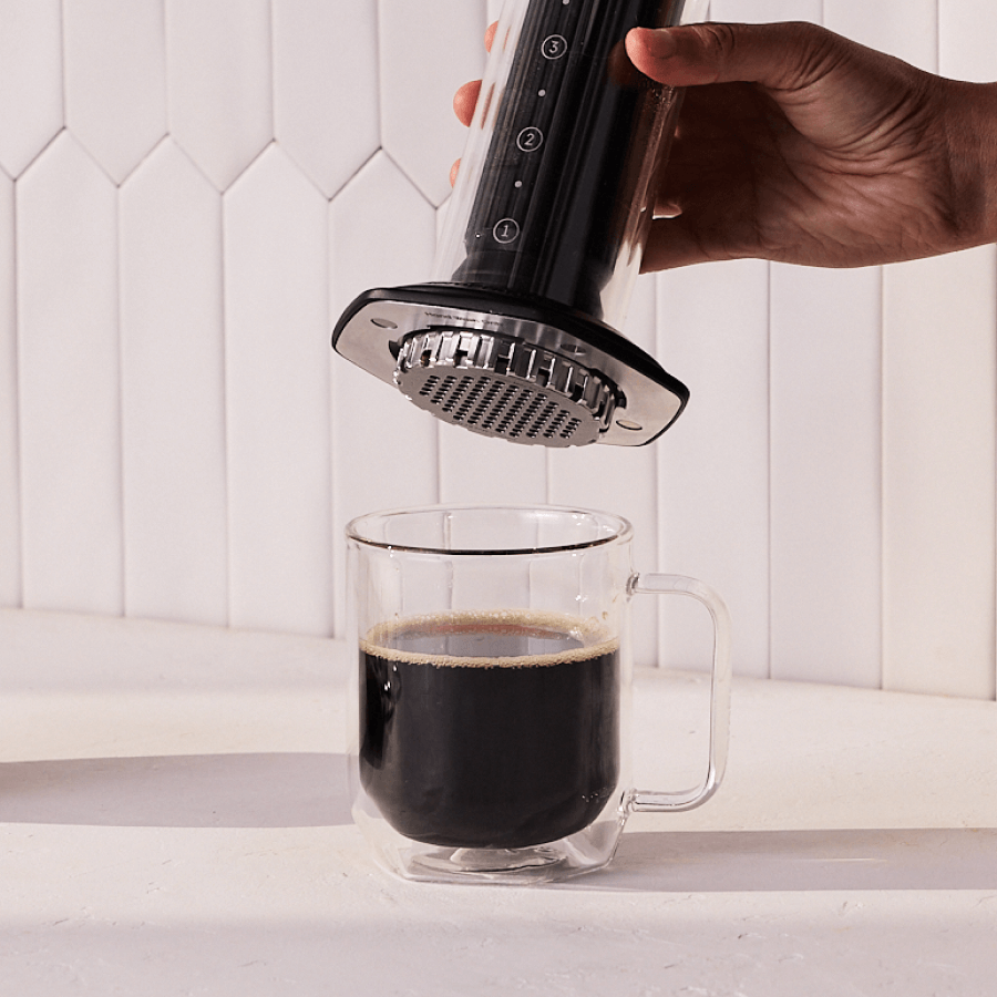 Hand using a AeroPress Premium coffee maker to pour coffee into a glass mug against a white tiled wall. #color_black
