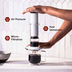 Person using a white AeroPress Premium coffee maker with text labels for air pressure, fast immersion, and micro-filtration. #color_white
