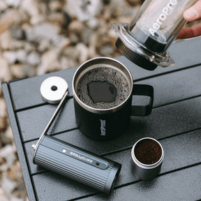 Aeropress coffee maker being used to pour coffee into a black mug on a wooden surface. #color_Purple