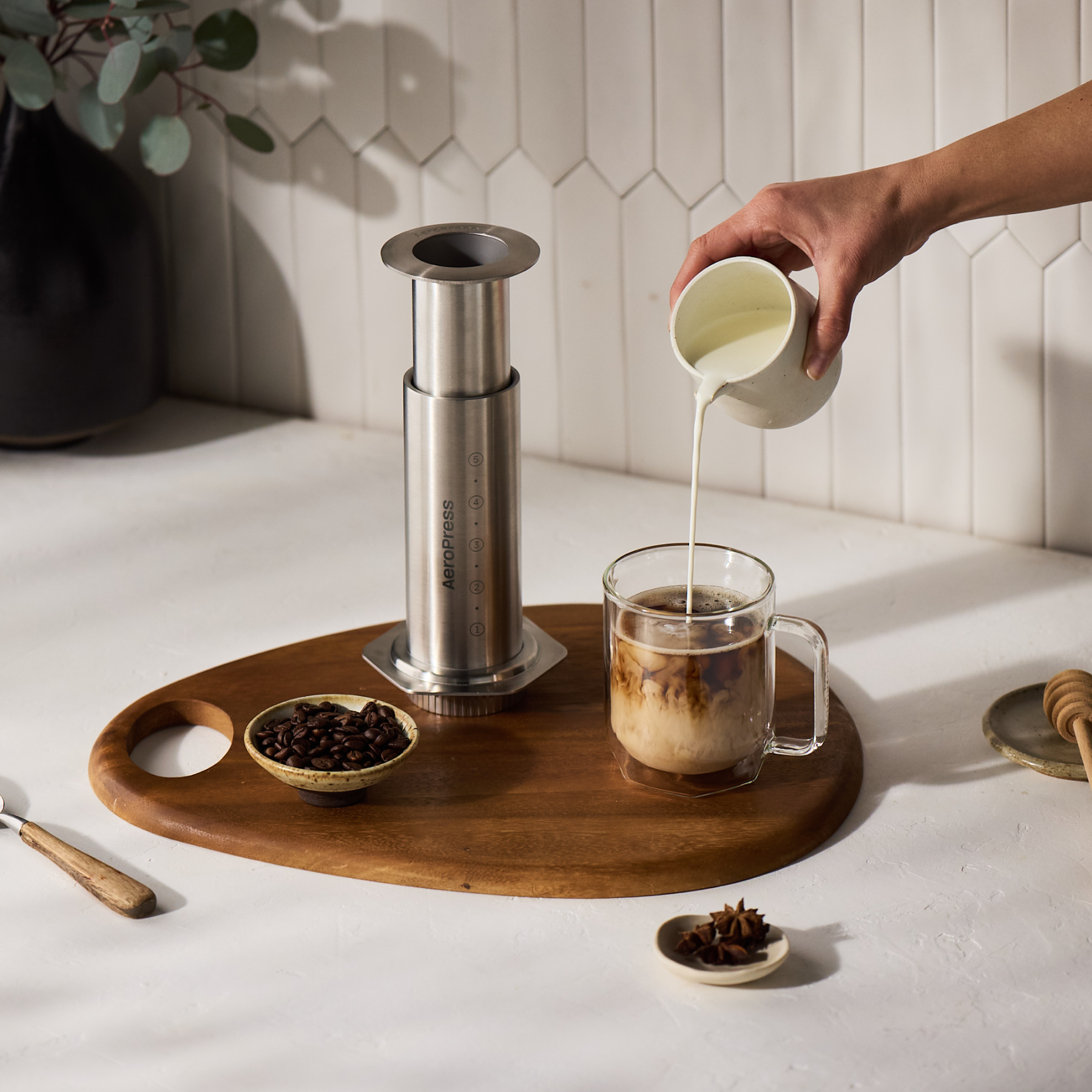 Person pouring milk into a coffee cup on a wooden tray with a AeroPress Steel coffee maker and coffee beans. #color_silver