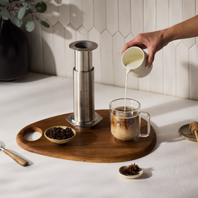 Person pouring milk into a coffee cup on a wooden tray with a AeroPress Steel coffee maker and coffee beans. #color_silver