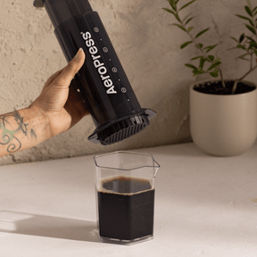 A person using an Aeropress to make coffee, with a glass of coffee on a table.