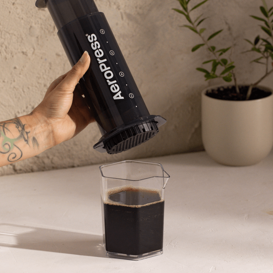 A person using an Aeropress to make coffee, with a glass of coffee on a table.