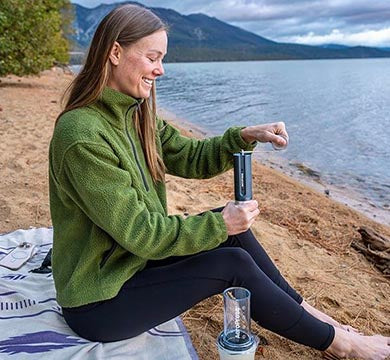 Woman sitting by a lake, grinding coffee by hand with a manual coffee grinder.