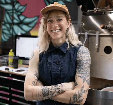 Person with tattoos standing in a coffee roastery with a coffee machine and computer in the background.