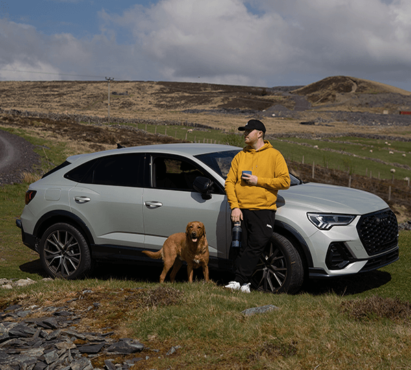 Person in yellow hoodie standing next to a silver SUV with a dog in a rural landscape