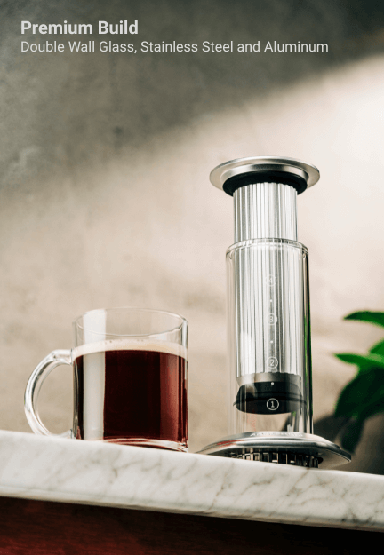 AeroPress-style coffee brewer constructed of double-wall glass, stainless steel, and aluminum, shown alongside a glass mug of dark coffee on a marble tray.